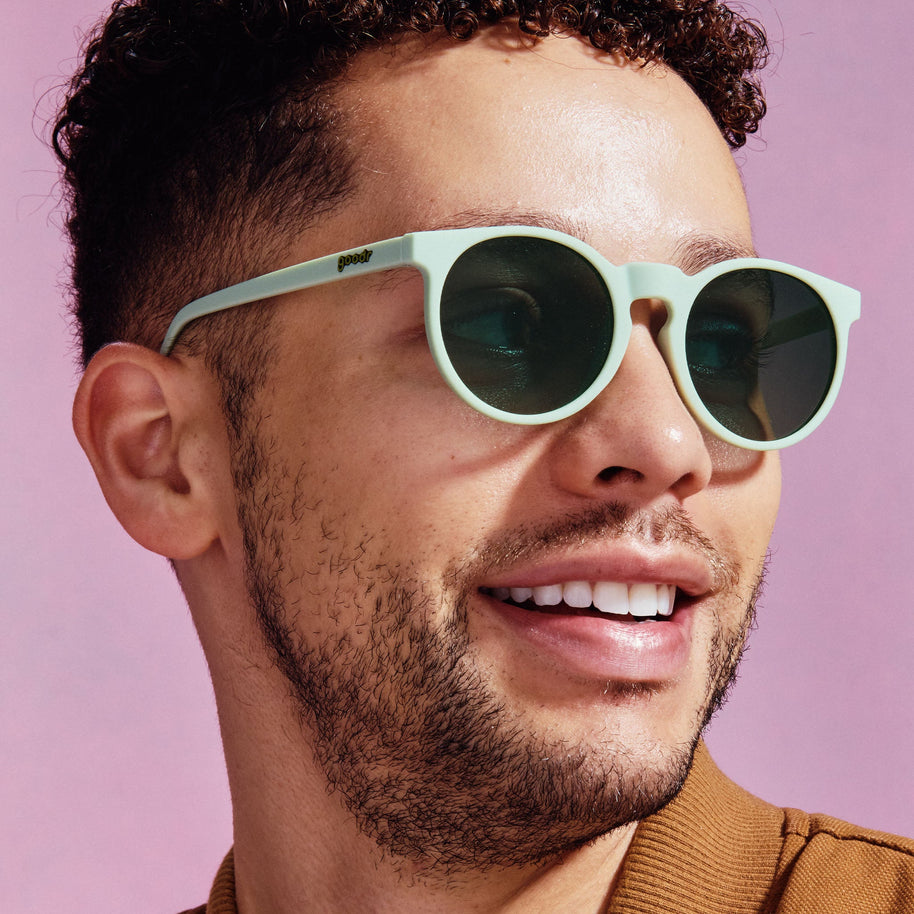 Smiling man against a pink background wearing Goodr Incognito Plant Parent sage green round sunglasses