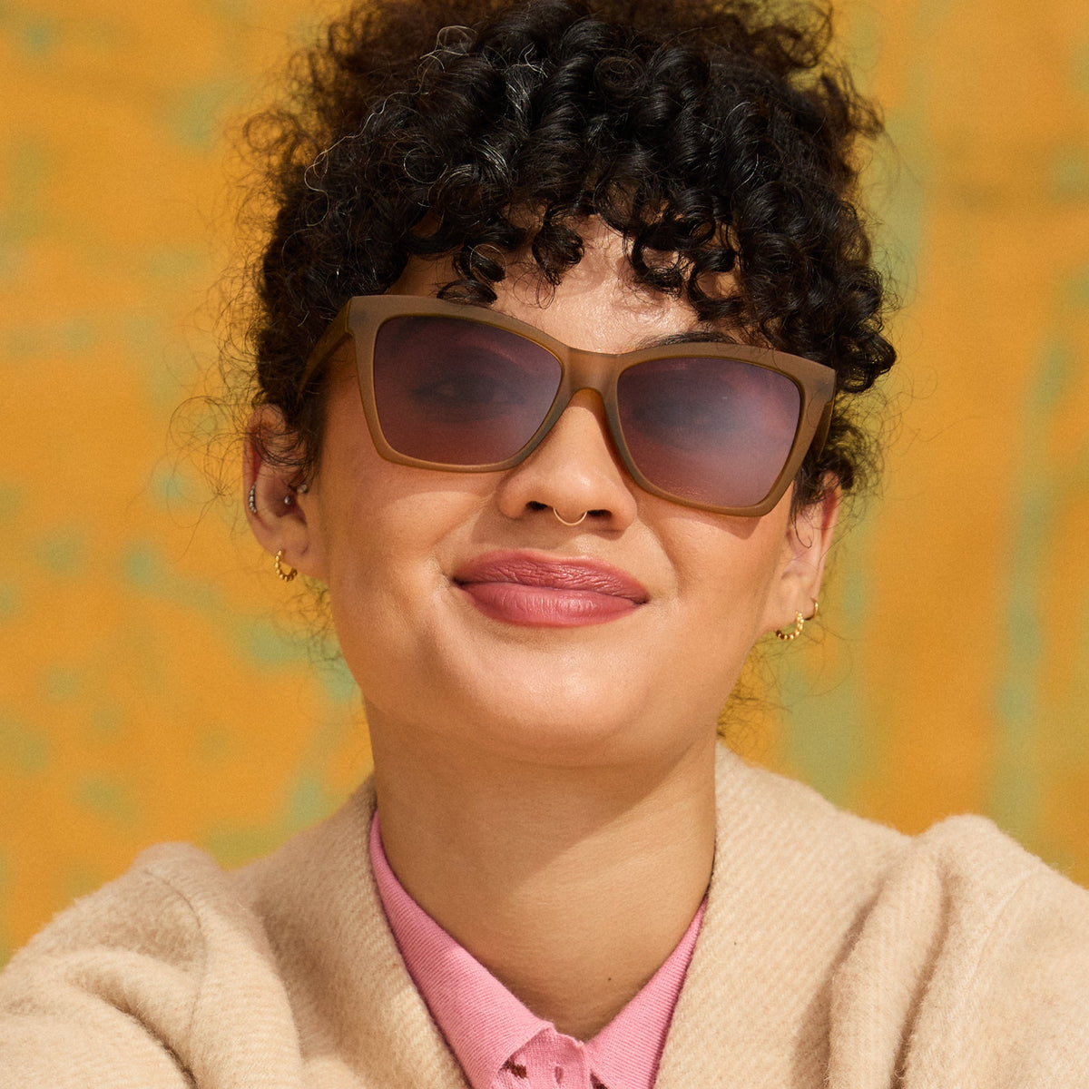 Smiling woman with curly hair in a cream cardigan wearing Goodr Sunbaked Sorcery sunglasses against a yellow wall
