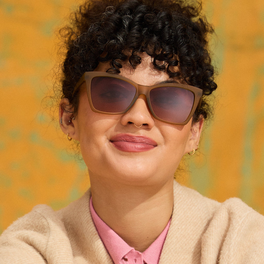 Smiling woman with curly hair in a cream cardigan wearing Goodr Sunbaked Sorcery sunglasses against a yellow wall
