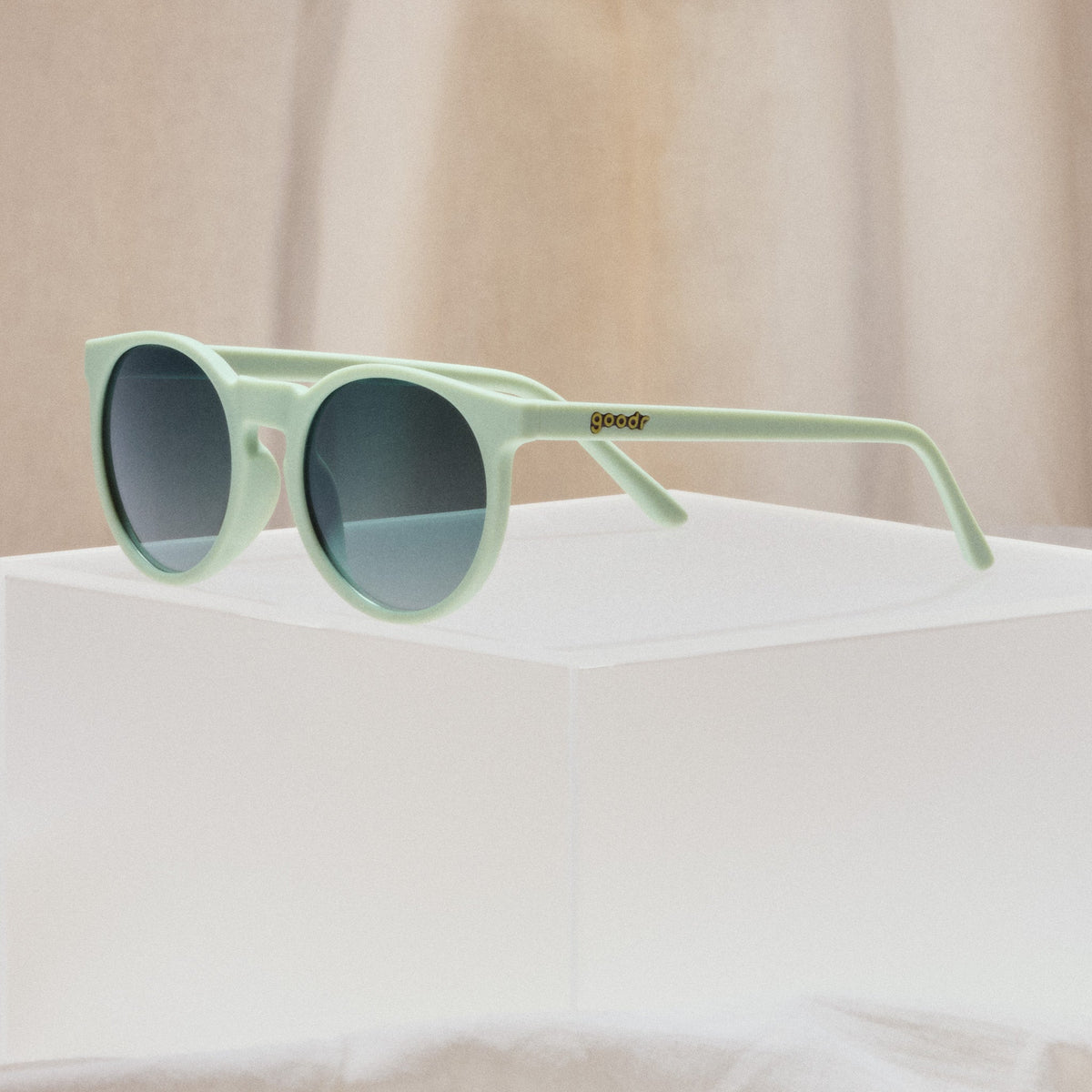 Goodr Incognito Plant Parent sunglasses resting on a white block in front of a beige draped backdrop
