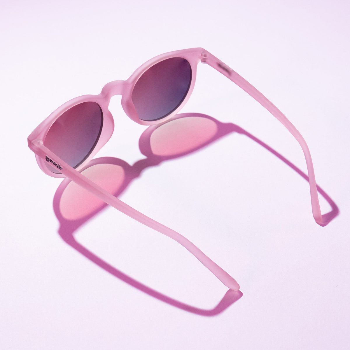 Goodr Mauve Mood Board sunglasses lying folded open on a light pink surface casting a pink shadow