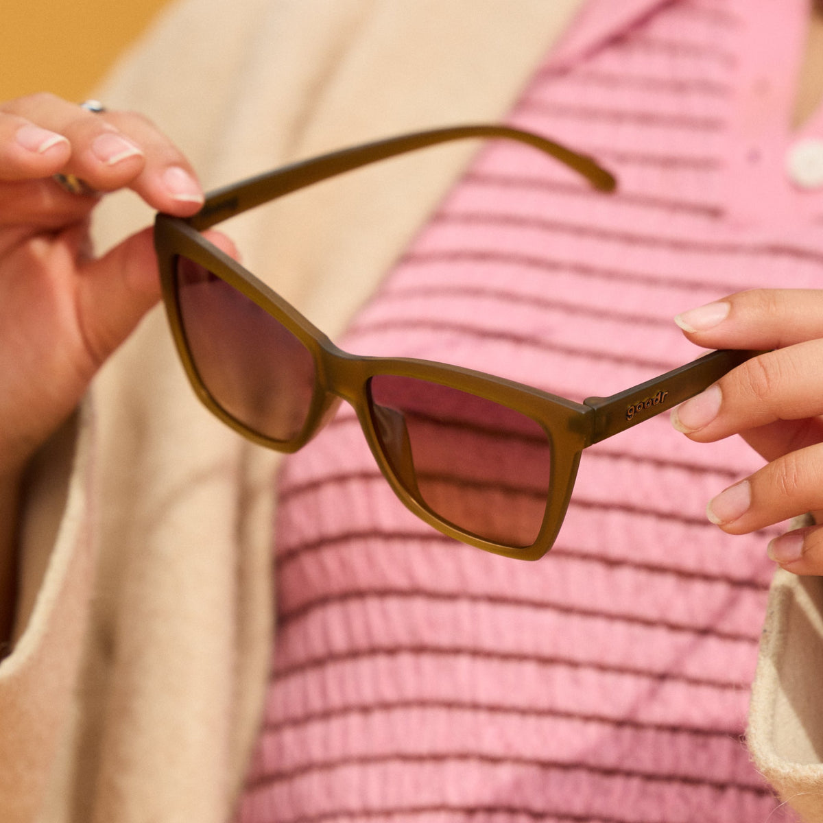 Hands holding Goodr Sunbaked Sorcery olive sunglasses in front of a pink striped top