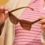 Hands holding Goodr Sunbaked Sorcery olive sunglasses in front of a pink striped top
