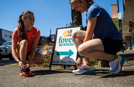 Two women tying shoelaces on lightweight running shoes in bright colors outdoors near a running store sign