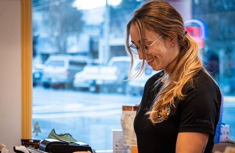 Young woman in black Craft sportswear with ponytail smiling indoors near window and electronic payment terminal