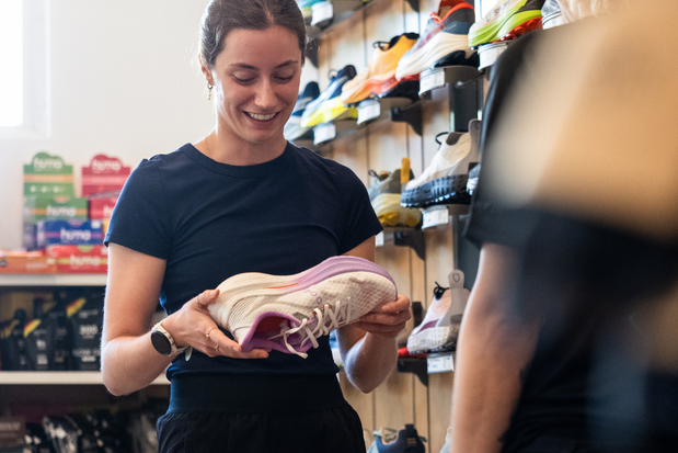 Woman in black shirt examining light pink and white running shoe inside a running store with wall display of various sports shoes