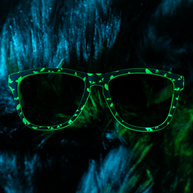 Front view of the Goodr What Lurks In The Shadows sunglasses with glowing green print on dark blue fur