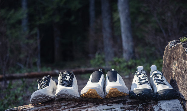 Three pairs of trail running shoes in beige, black, and grey on a log in forest setting for outdoor running