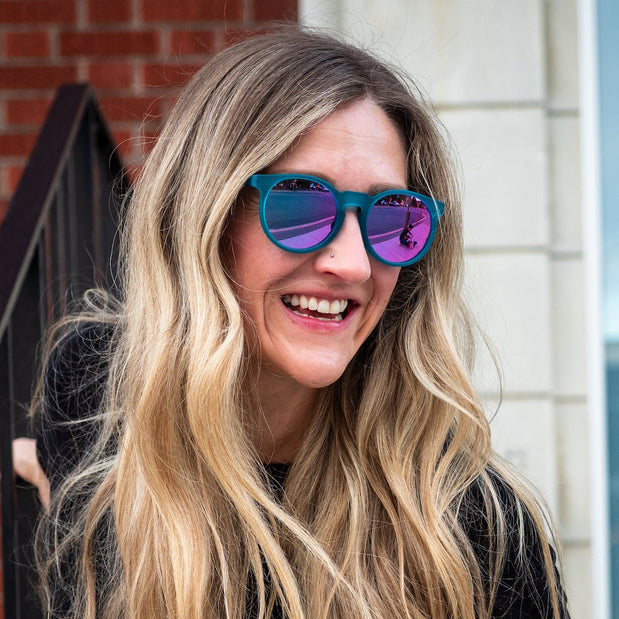 Woman with long blonde hair wearing teal frame sunglasses with purple reflective lenses smiling outdoors by building stairs