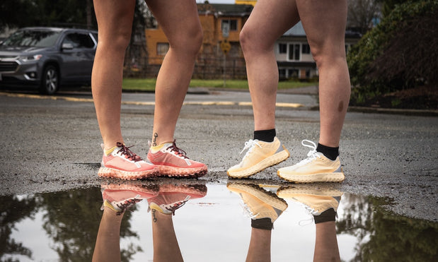 Two people standing on wet pavement wearing peach and cream lightweight running shoes with rugged outsoles reflected in a puddle