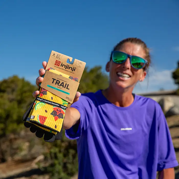 Smiling woman in sunglasses holding up a packaged pair of Injinji The Courtney Crew toe socks in Solar Slice