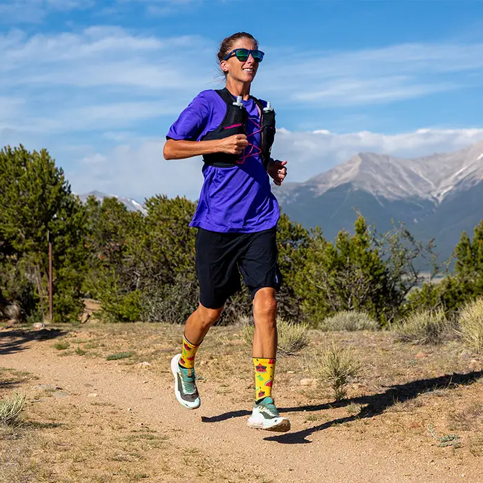 Trail runner in a purple shirt and vest on a dirt path below mountains wearing Injinji The Courtney Crew socks in Solar Slice
