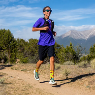Trail runner in a purple shirt and vest on a dirt path below mountains wearing Injinji The Courtney Crew socks in Solar Slice