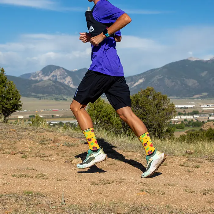 Runner striding along a dry dirt trail with mountains behind, wearing yellow Injinji The Courtney Crew socks in Solar Slice