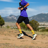 Runner striding along a dry dirt trail with mountains behind, wearing yellow Injinji The Courtney Crew socks in Solar Slice