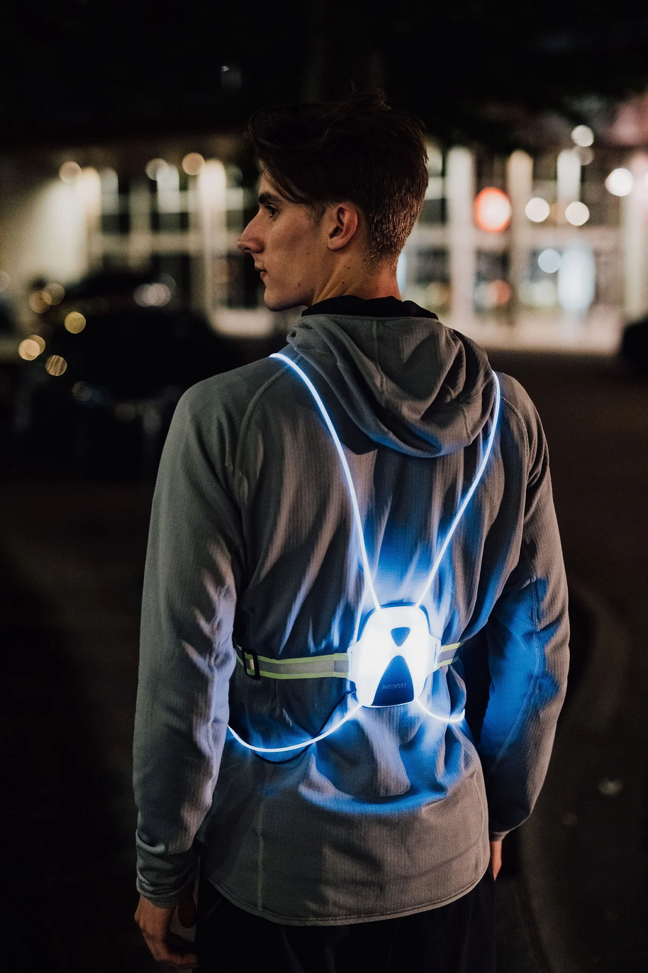 Runner at night on a city street, back view wearing a NiteVest Rechargeable LED Safety Vest glowing blue over a grey hoodie