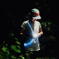 Runner in a cap on a dark trail among plants wearing a glowing blue NiteVest Rechargeable LED Safety Vest