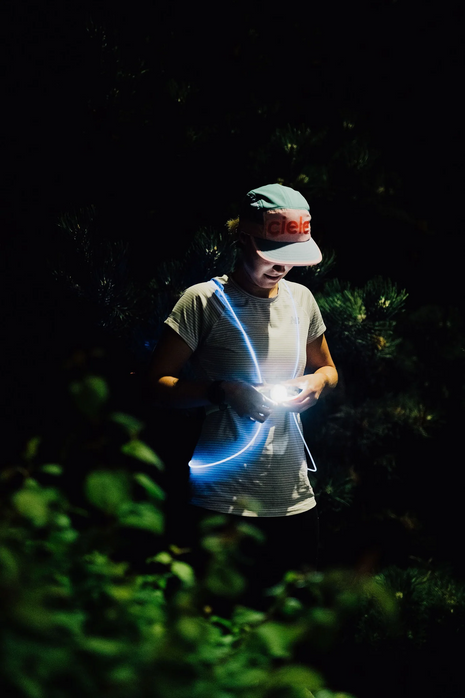 Runner in a cap on a dark trail among plants wearing a glowing blue NiteVest Rechargeable LED Safety Vest