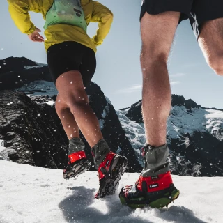Runners on snow below mountain peaks wearing Kahtoola LEVAgaiter GTX Mid gaiters in red and grey