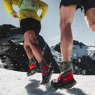 Runners on snow below mountain peaks wearing Kahtoola LEVAgaiter GTX Mid gaiters in red and grey