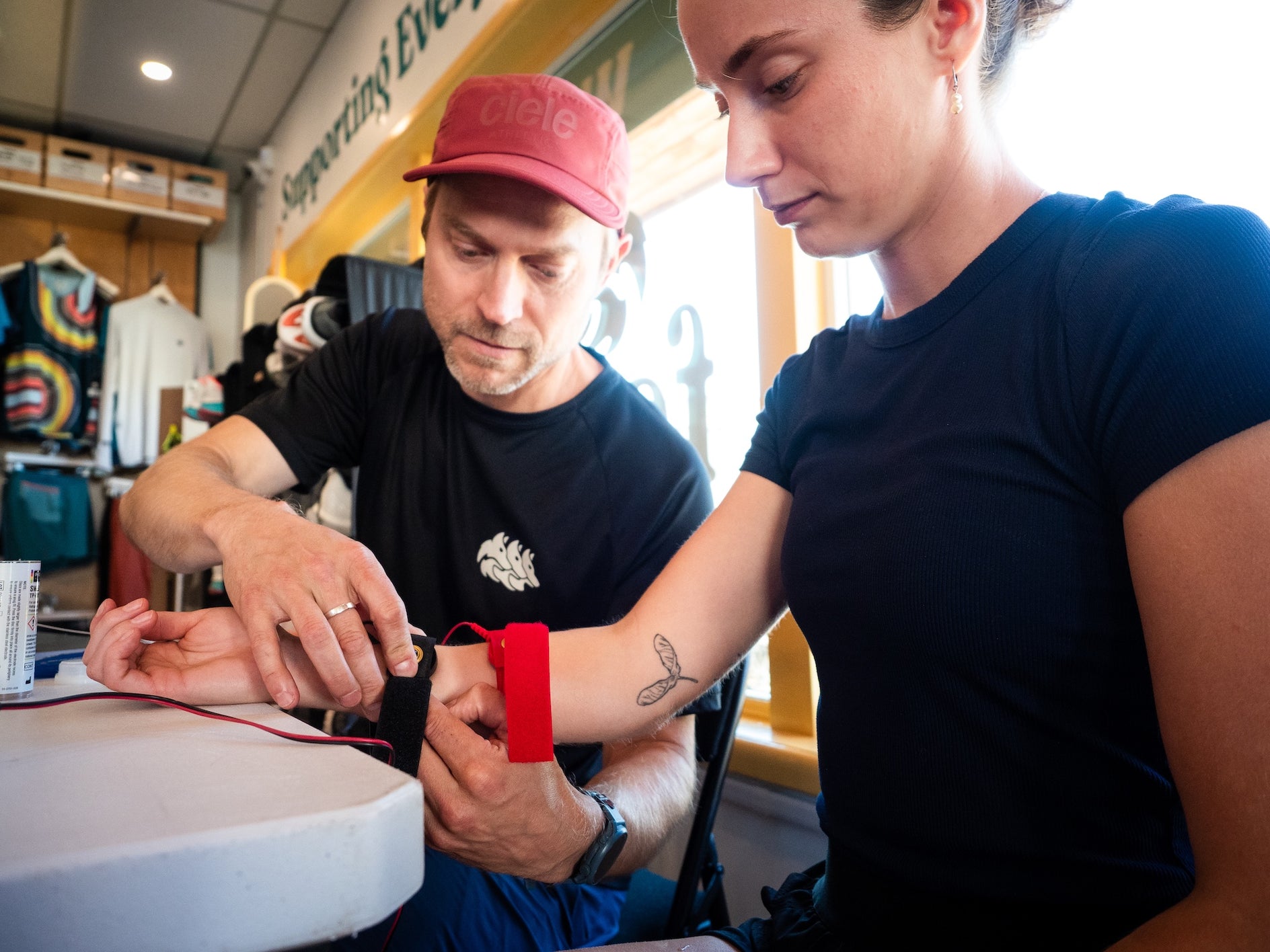Man in red cap adjusting heart rate monitor strap on woman in black shirt at indoor sports shop table