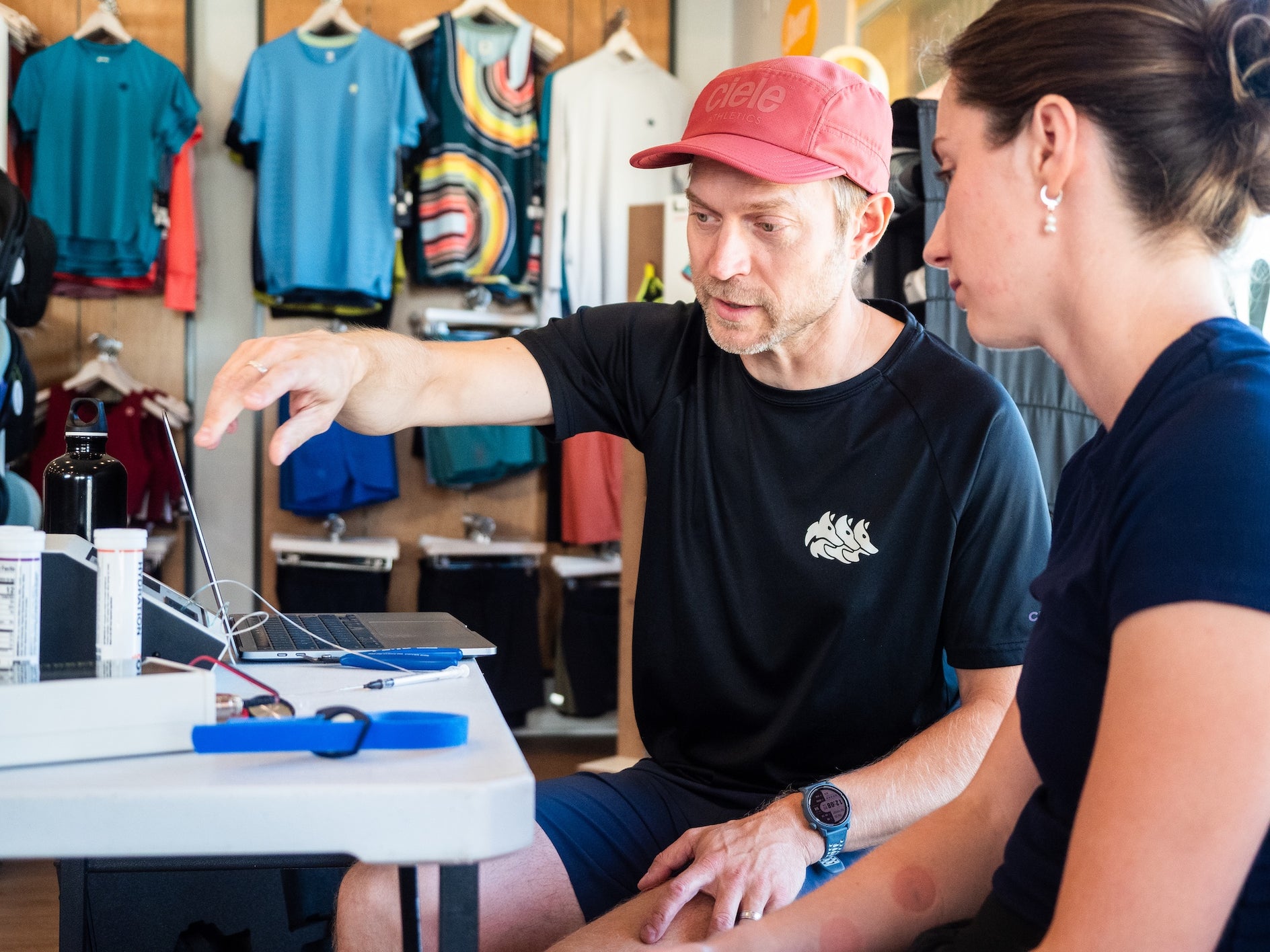 Man in pink cap and black t-shirt explaining running gear details to woman in navy shirt in a running store setting